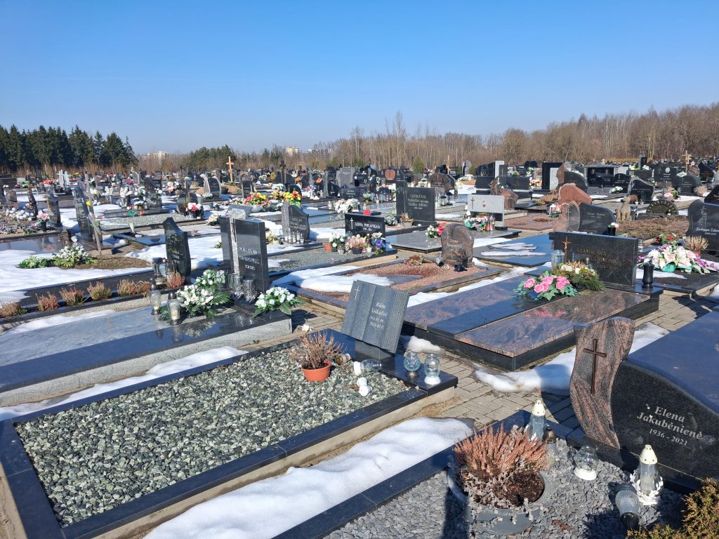 20260309_114149 - Yoshin matkassa Wide cemetery scene with rows of gravestones, flowers, and candles, snow on the ground under a clear blue sky.