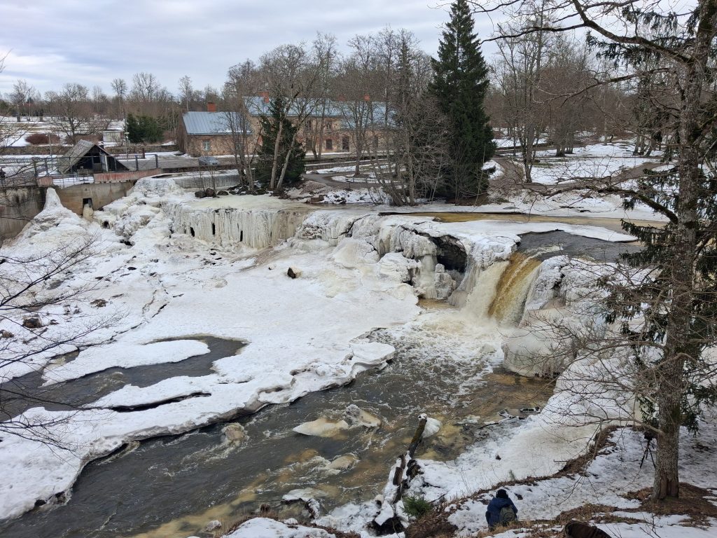 Snowy riverside with a frozen waterfall and ice formations, a brick building in the background, and a person in the lower-right corner.