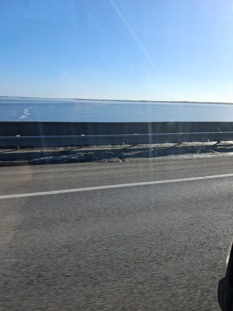 Guardrail along a highway beside a frozen lake under a clear blue sky; photo taken from a moving car.